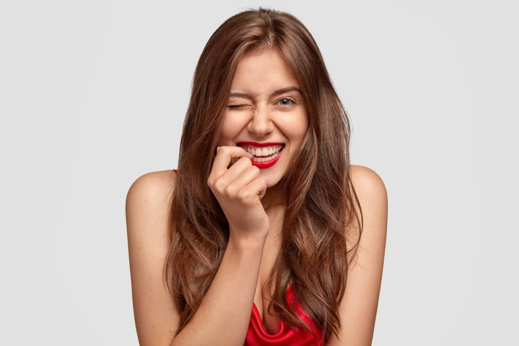 Hollywood smile longevity example—winking woman with bright white veneer smile and red lipstick on a neutral background.